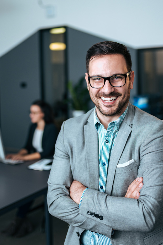 In the foreground is a middle aged man with glasses in a suit with an unbuttoned collar and no tie. He's smiling. In the background is an office where a woman is working on a computer.